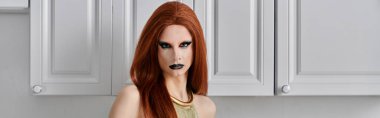 Bir travesti mutfakta beyaz dolaplarla poz veriyor, göz kamaştırıcı bir kıyafet giyiyor..