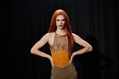 Işıldayan altın korseli ve uzun kızıl saçlı bir travesti siyah bir arkaplan önünde güven içinde poz verir..