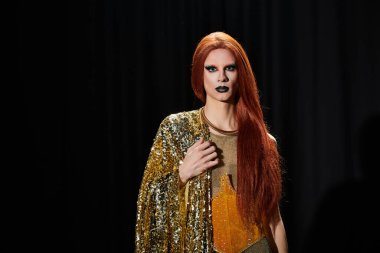 Bir travesti, göz kamaştırıcı bir altın elbiseyle karanlık bir arkaplanda poz verir..