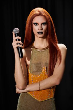 Bir travesti mikrofonla poz verir, üzerinde altın ve turuncu bir kıyafet vardır..