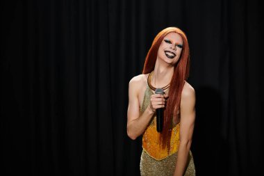 Altın pullu elbiseli bir travesti elinde mikrofon tutarken gülümsüyor..