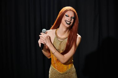 Bir travesti sahnede, elinde bir mikrofon, parlak bir gülümsemeyle şarkı söylüyor..