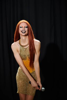 Işıldayan altın elbiseli bir drag sanatçısı bir mikrofon tutuyor, eğlendirmeye hazır..