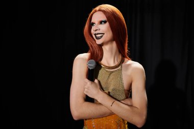 Kızıl saçlı ve kalın makyajlı bir travesti mikrofonla poz veriyor..