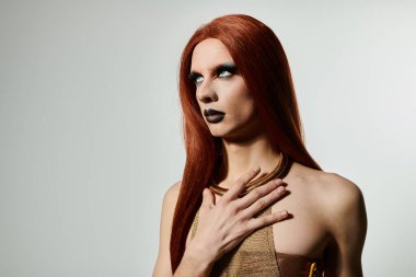 Uzun kızıl saçlı ve dramatik makyajlı bir drag diva yukarı bakıyor, eli göğsünde dinleniyor..