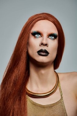Uzun kızıl saçlı, dramatik makyajlı, altın bir bluz ve kolye takan bir travesti..