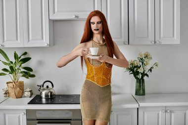 Parlak elbiseli bir travesti, el değmemiş bir mutfakta bir fincan çayın tadını çıkarıyor..