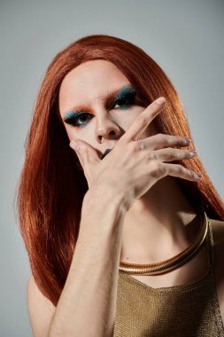 Kızıl saçlı ve dramatik makyajlı bir travesti, ağzı açık, altın pullu bir bluz ve boğucu kolye takıyor..