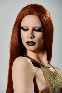 Uzun kızıl saçlı ve kalın makyajlı bir travesti kameraya kendinden emin bir şekilde bakar..
