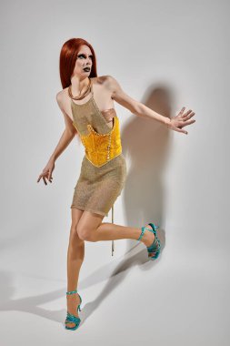 Büyüleyici altın elbiseli bir travesti beyaz bir duvara karşı poz verir..