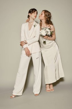 Beyaz düğün kıyafeti giymiş iki kadın, gri bir zemine karşı birlikte poz veriyorlar, LGBTQ + sevgi ve bağlılığı simgeliyorlar..