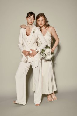 Two women in white attire stand together for their wedding. One wears a suit, while the other is in a dress. clipart