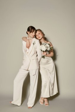 Two young women in white wedding attire pose against a gray background, celebrating their love. clipart