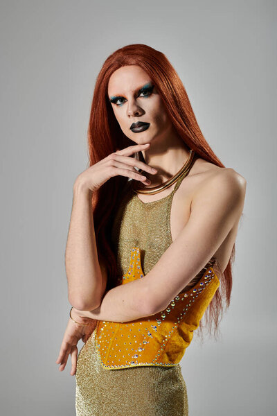 A drag queen in a gold sequined dress and sparkling makeup poses in a studio.