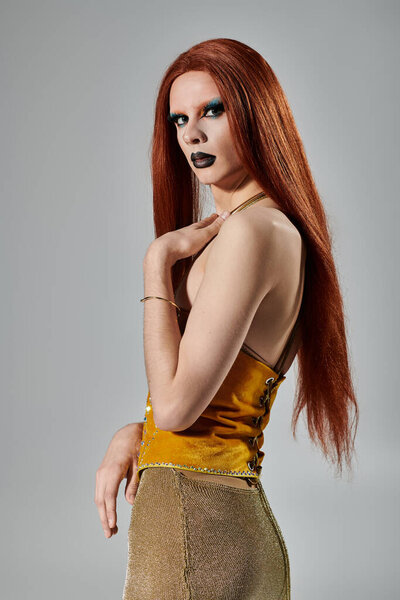 A drag queen with long red hair and bold makeup poses in a glamorous gold outfit.