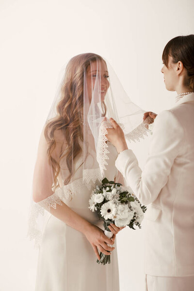 A bride in a white dress adjusts her veil as her partner stands behind her, ready to celebrate their special day.