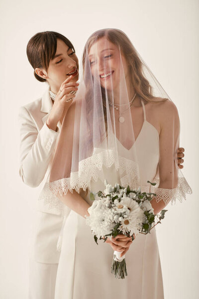 Two brides in white wedding attire, one holding a bouquet of white flowers.
