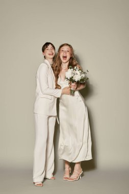 Beyaz elbiseli lezbiyen bir çift, gri arka planda yakalanarak düğün günlerini kutluyor. Her ikisi de poz verirken ışıl ışıl gülümserler..