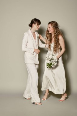 A lesbian couple in white attire smiles at each other during their wedding celebration, posing against a grey background. clipart