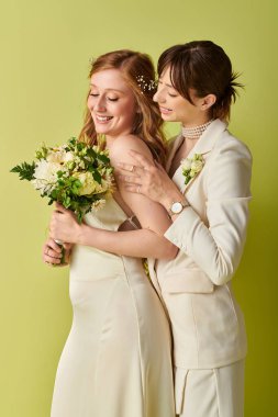 Beyaz düğün kıyafetli iki gelin birbirine sarılıyor, yeşil bir zeminde..