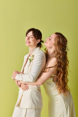Beyaz düğün kıyafetli iki kadın yeşil bir zemine karşı sarılıyorlar..