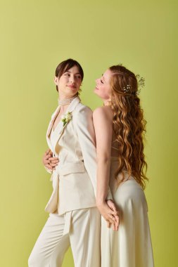 Beyaz düğün kıyafetli iki kadın yan yana duruyor, gülümsüyor ve yeşil arka planda birbirlerine bakıyorlar..