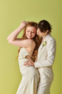Beyaz düğün kıyafetli iki kadın yeşil arka planda sarılıyor. Çift gülümsüyor ve birbirine sevgiyle bakıyor..