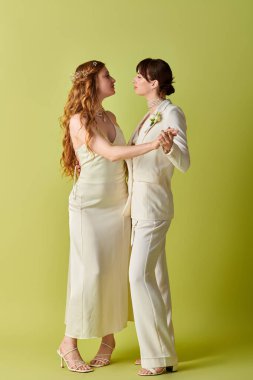 Beyaz düğün kıyafetli iki gelin, birbirine sevgiyle bakıyor, canlı yeşil bir arka planda..