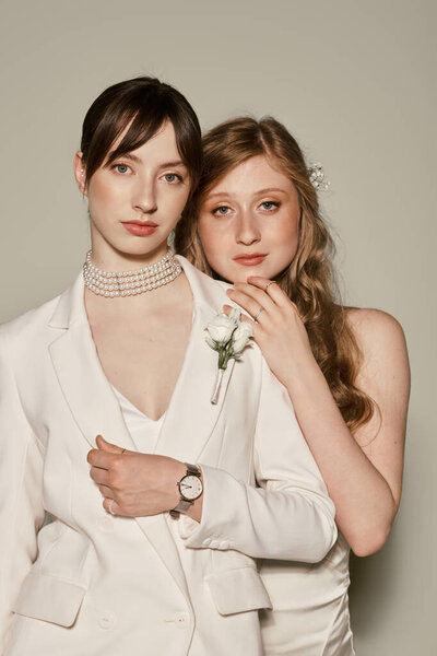 Две молодые женщины в белой свадебной одежде обнимаются на сером фоне.