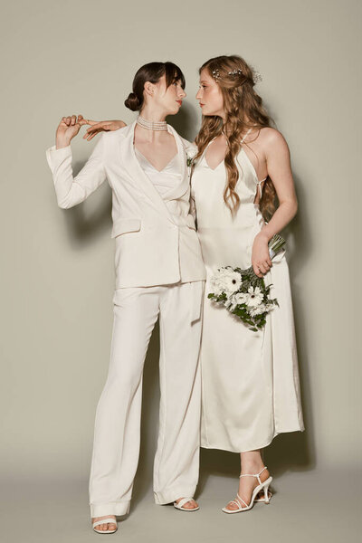 Two women in white attire, one wearing a suit and the other a dress, embrace during their wedding ceremony.