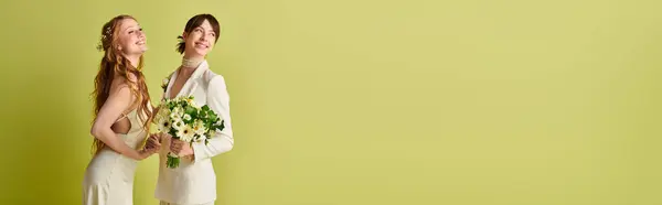 stock image Two women in white wedding attire stand against a green background. One holds a bouquet of flowers while the other smiles happily.