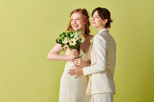 Two women in white wedding attire embrace, laughing with joy on a green backdrop. One holds a bouquet of white flowers.