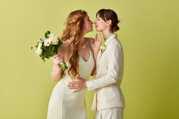 Две невесты в белом разделяют нежный поцелуй во время их свадебной церемонии, их радость ощущается на зеленом фоне.