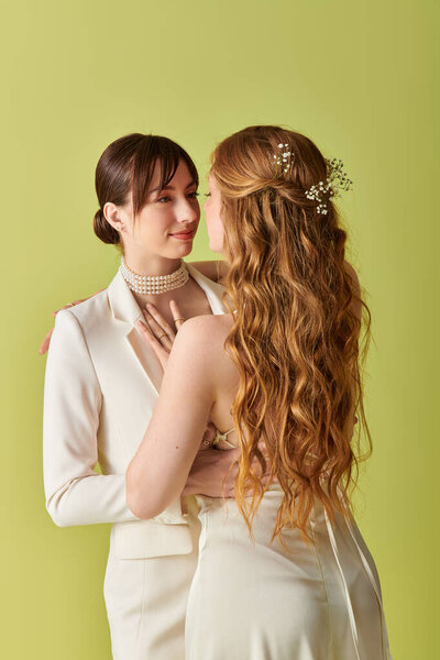 A young lesbian couple in white attire embrace during their wedding ceremony, their love evident in their gaze.
