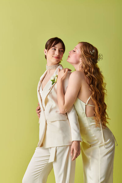 A lesbian couple, dressed in white, embrace during their wedding ceremony. They stand against a bright green background, radiating joy and love.