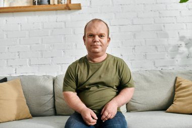 Kapsamlı bir adam kanepeye oturur ve sakince kameraya bakar..