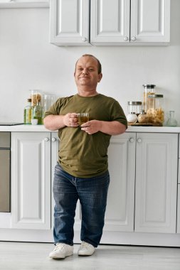 Kapsayıcı bir adam elinde bir bardak içecekle mutfağında durur ve gülümser..