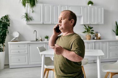 A man with inclusivity wearing casual attire stands in a kitchen and speaks on a phone. clipart