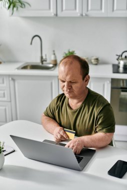 Kapsamlı bir adam mutfak masasında dizüstü bilgisayarını ve kredi kartını online alışveriş için kullanır..