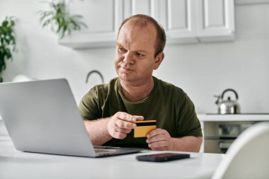 Kapsamlı bir adam mutfak masasında oturmuş kredi kartıyla online alışveriş yapıyor..
