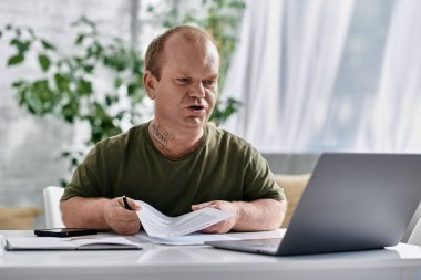 A man with inclusivity sits at a desk in a home office, reviewing documents while looking at a laptop. clipart