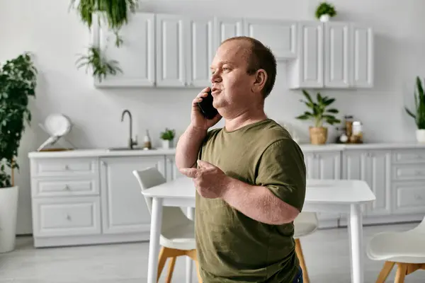 stock image A man with inclusivity wearing casual attire stands in a kitchen and speaks on a phone.