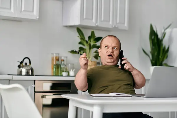 stock image A man with inclusivity sits at a kitchen table, talking on the phone, with a laptop in front of him.