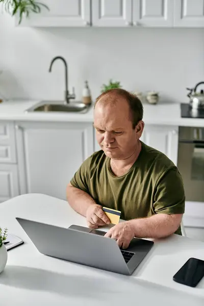 stock image A man with inclusivity sits at a kitchen table, using his laptop and a credit card for online shopping.