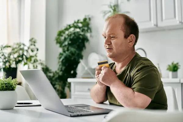 stock image A man with inclusivity is sitting at a table in his home, using a laptop to make a purchase online. He is holding a credit card in his hand.