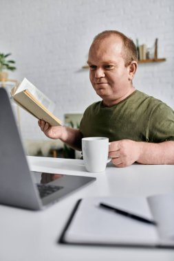 Günlük kıyafetlerle dolu bir adam evinde bir masaya oturur, kitap okur ve bir fincan kahvenin tadını çıkarır..