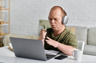 Günlük kıyafetleri olan bir adam bir masaya oturur, kulaklık takar ve dizüstü bilgisayar kullanır..