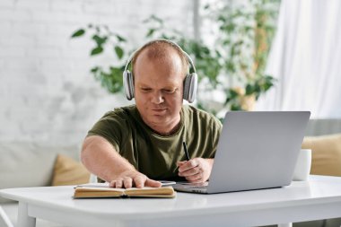 Kapsayıcı kulaklık takan bir adam elinde dizüstü bilgisayar ve kitapla bir masada oturup evde ders çalışıyor..