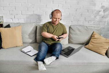 A man with inclusivity relaxes on a couch at home, wearing headphones and checking his phone. clipart