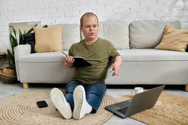 Kapsamlı bir adam koltuğun önünde yerde oturuyor ve yanında dizüstü bilgisayarı olan bir defter üzerinde çalışıyor..